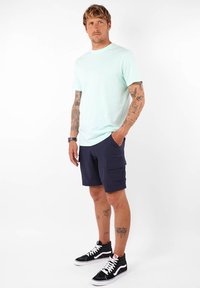 T-shirt vert menthe à col rond, shorts cargo bleu marine avec poches latérales, et baskets montantes noires avec accents blancs, portés par un homme.