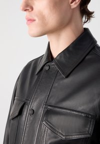 Μαύρο δερμάτινο μπουφάν με γιακά, διαθέτοντας δύο μπροστινές τσέπες, κλείσιμο με κουμπιά και εμφανή διακοσμητικά ραφές κατά μήκος των ραφών.