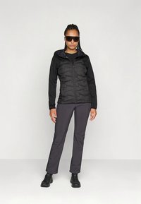 Melna izolēta jaka ar rāvējslēdzēju priekšpusē un kapuci, kopā ar tumši pelēkām biksēm un melniem apaviem, ar plānu, pieguļošu dizainu.