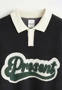 Polo noir avec col et boutons blancs, présentant un lettrage texturé vert et blanc "Présent" sur la poitrine.