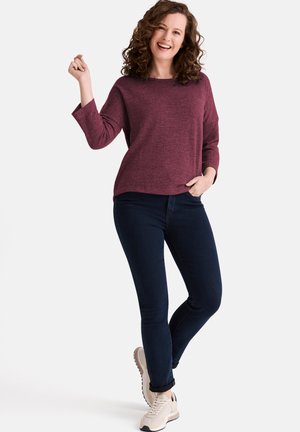 Weinrotes Strickoberteil mit langen Ärmeln, lockerer Passform, kombiniert mit dunkelblauen Skinny-Jeans und hellen Turnschuhen. Das Model posiert mit erhobener Hand.
