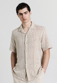 Chemise à manches courtes beige avec un motif en zigzag, présentant un devant boutonné, un col évasé et deux poches avant. Tissu texturé.