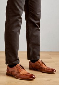 Hnědé kožené společenské boty s brogue detaily, kulatou špičkou a šněrováním, v kombinaci s tmavorůžovými kalhotami na dřevěné podlaze.