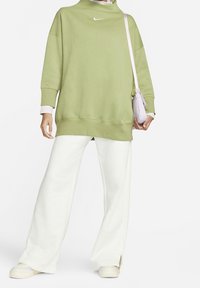 Grön sweatshirt med hög krage, vit logotyp, matchad med vita vida byxor och ljust färgade skor, bärandes en liten lavendelväska.