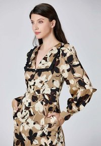Robe à imprimé floral beige avec des accents noirs, présentant des manches bouffantes, un décolleté en V profond et des poches avant. Détails avec des volants.