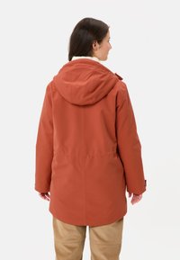 Giacca impermeabile di lunghezza media di colore ruggine, con cappuccio, fodera morbida, polsini regolabili e vita con coulisse. Texture liscia e taglio dritto.