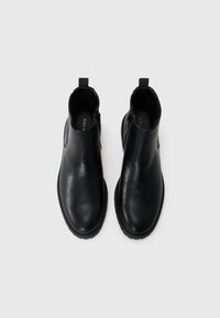 Botas de tobillo de cuero negro con una textura suave, puntera redondeada, paneles laterales elásticos y tiradores para facilitar el uso. Suelas de goma para mayor agarre.