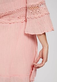 Vestido de un rosa claro, texturizado, con un delicado encaje en las mangas y la cintura. Mangas con bordes acampanados y con volantes, tejido suave con pliegues visibles.