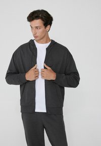 Sudadera gris con cremallera hecha de tejido suave, que cuenta con capucha ajustable y bolsillos frontales, llevada encima de una camisa blanca. El modelo se muestra de manera casual.