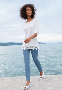 Witte lange mouwen top met kantafwerking, in combinatie met lichtblauwe broek en witte sandalen. Model staat bij water tegen een bewolkte lucht.