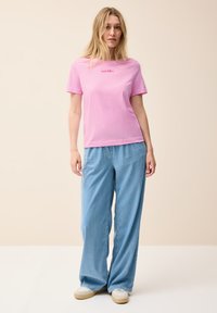 Camiseta de algodón rosa con mangas cortas que presenta un pequeño logo. Combinada con vaqueros de pierna ancha de color azul claro y zapatillas beige. Diseño sencillo.