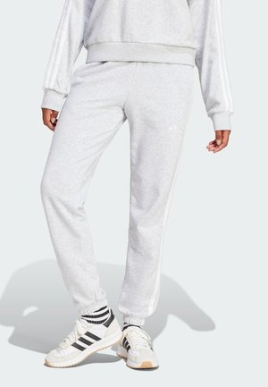 Person wearing light gray Adidas sweatpants and matching sweatshirt with white and black Adidas sneakers, standing against a light background.