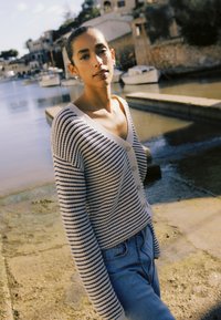 Ung kvinde iført stribet cardigan og jeans står ved en rolig havn med fortøjede både og stenhuse i baggrunden.