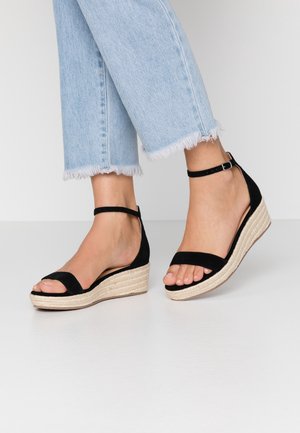 Füße in schwarzen Keilsandalen mit Knöchelriemen, geflochtenen Sohlen und hellblauen Jeans mit ausgefranstem Saum auf weißem Hintergrund.