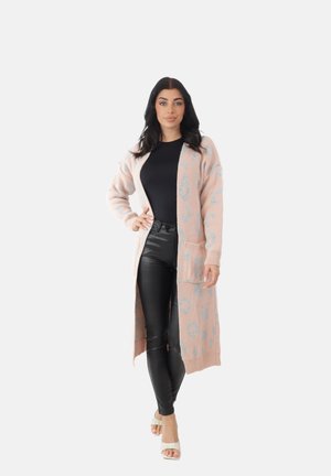 Femme debout, main sur la hanche, portant un long cardigan rose à motifs, un haut noir, un pantalon en cuir noir et des sandales blanches à talons.