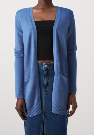 Femme portant un cardigan bleu clair ouvert sur un crop top noir et une jupe en jean taille haute fendue à l'avant.