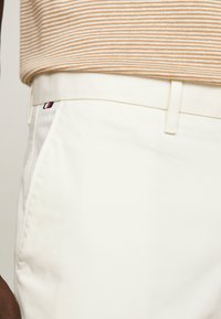 Pantaloni bianchi in cotone con una vestibilità su misura, dotati di tasche laterali e una piccola patch con logo rosso, bianco e blu vicino alla vita.