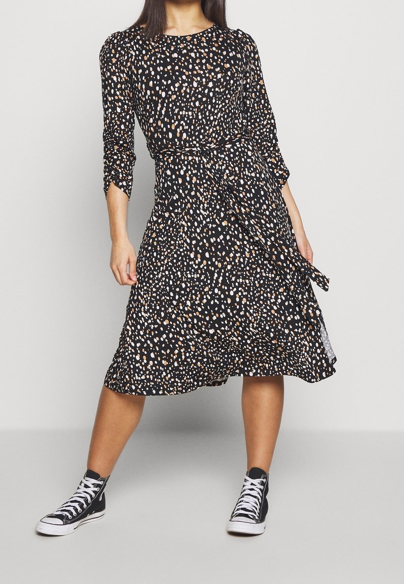 Frau in einem knielangen Kleid mit schwarz-weißen und braunen Flecken, mit Dreiviertelärmel und schwarzen High-Top-Sneakern, die mit gespreizten Beinen steht.