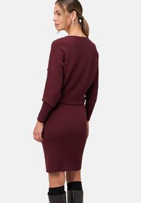 Robe en tricot côtelé bordeaux avec des manches longues, forme ajustée et taille accentuée. Portée avec des bottes noires brillantes jusqu'aux genoux.
