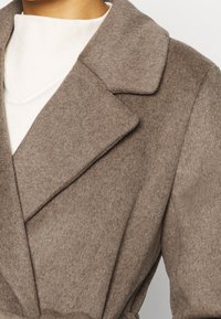 Bruin wollen jas met een brede kraag, een ceintuur in de taille en een gestructureerde stof. V-hals witte top zichtbaar eronder.