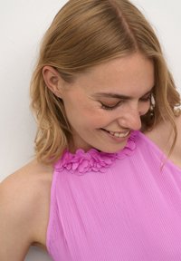 Nuori nainen, jolla on vaaleanruskeat hiukset, on pukeutunut vaaleanpunaiseen olkaimettomaan puseroon, jossa on kukkakoristeita. Hän hymyilee ja katsoo alas.