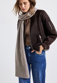 Kvinna iklädd en brun bomberjacka, blå jeans, brun topp och lång gr�å scarf, med en hand i jeansfickan.