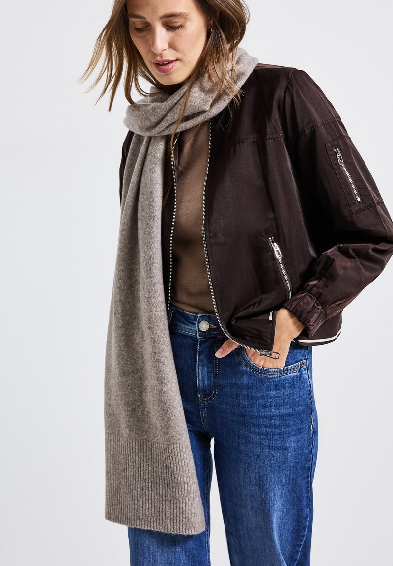 Kvinna iklädd en brun bomberjacka, blå jeans, brun topp och lång grå scarf, med en hand i jeansfickan.