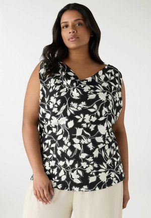 Donna che indossa una canotta nera con stampa floreale bianca e pantaloni beige, in piedi contro uno sfondo chiaro e semplice.