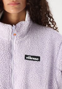 Lavendel fleecejakke med høy krage, glidelås og svart "ellesse" logo-patch på fronten. Teksturert, plysj-lerret.