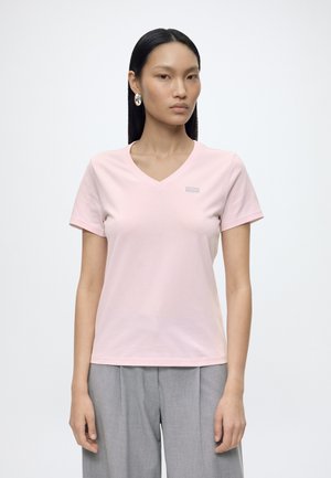 Femme aux cheveux noirs et raides porte un t-shirt rose pâle à col en V et manches courtes, ainsi qu'un pantalon gris taille haute, debout devant un fond uni.