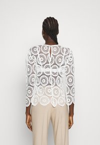 Haut en dentelle blanche avec des motifs floraux, des manches longues et un ourlet festonné. Le dos présente un petit détail en forme de clé et une texture transparente.