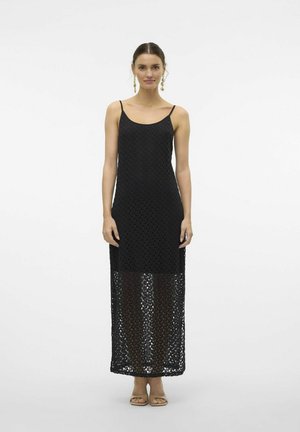 Abito lungo in crochet nero con spalline sottili, motivo a rete testurizzato e orlo trasparente. Indossato con sandali bassi neutri.