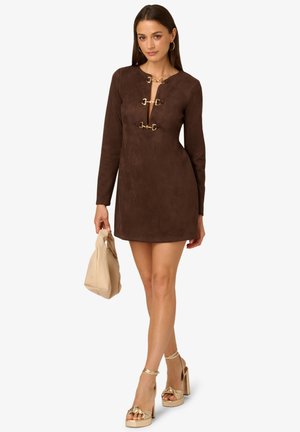 Bruine suede mini-jurk met lange mouwen, diepe halslijn en gouden hardware-accenten. Gecombineerd met een beige tas en gouden platformhakken.