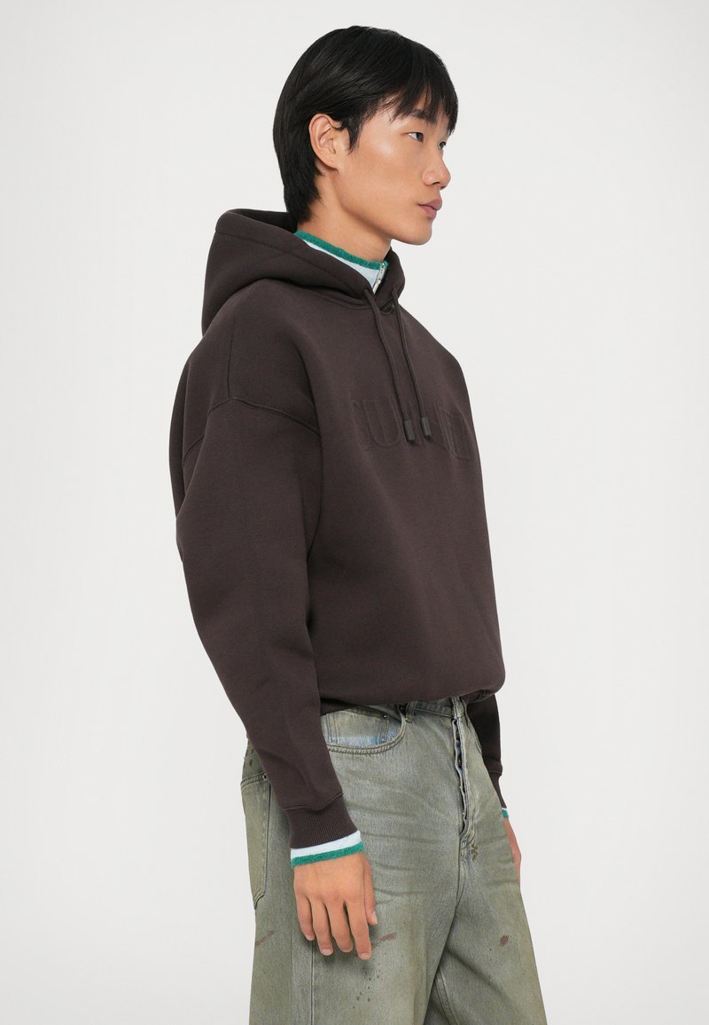 Felpa marrone con tasca a marsupio, cordini e logo in rilievo. Abbinate a jeans chiari in denim con un risvolto a strisce verdi e bianche.