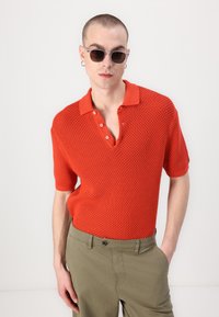 Rød strikket polo med teksturert mønster, korte ermer og krage med tre knapper. Kombinert med lyse olivengrønne bukser.