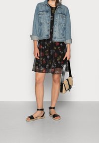 Veste en denim, robe fleurie noire avec des accents rouges et jaunes, et sandales noires avec une semelle texturée ; portant un petit sac en osier.