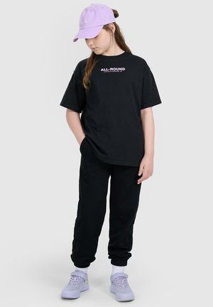 Dziewczyna ubrana w czarny oversize'owy T-shirt i spodnie, lawendową czapkę z daszkiem i pasujące trampki, stoi z jedną ręką w kieszeni, patrząc w dół.