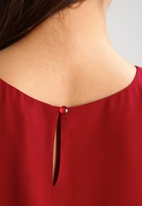 Gros plan sur le dos supérieur d'une femme portant un chemisier rouge avec une ouverture en forme de clé fermée par un petit bouton rouge.