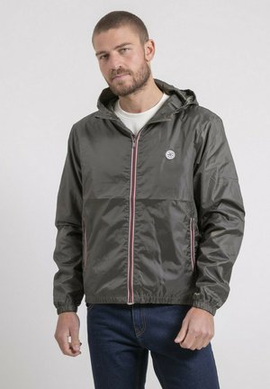 Homme portant une veste à capuche vert olive avec fermeture éclair et poches à l'avant, debout devant un fond uni clair.