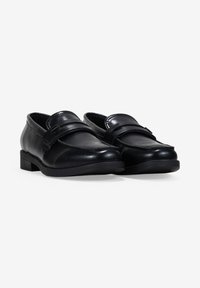 Zapatos de piel negra con puntera redonda, que cuentan con una tira decorativa en el empeine y un tacón bajo. Textura suave y detalles mínimos.