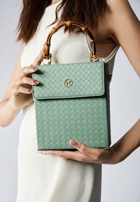 Zielona, pleciona torebka z teksturowym wykończeniem, z bambusowym uchwytem i złotym logo. Posiada zapięcie na klapkę oraz usztywniony prostokątny kształt.