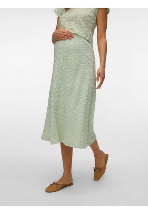 Femme enceinte portant une robe midi bleu clair à texture florale et des ballerines marron clair sans lacets, tenant son ventre d'une main.
