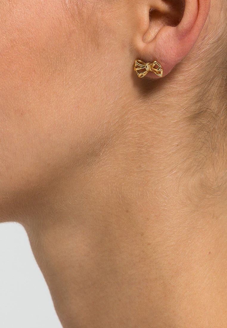 Gros plan d'une boucle d'oreille clou en forme de nœud en or portée sur le lobe gauche d'une personne à la peau claire et aux cheveux fins.