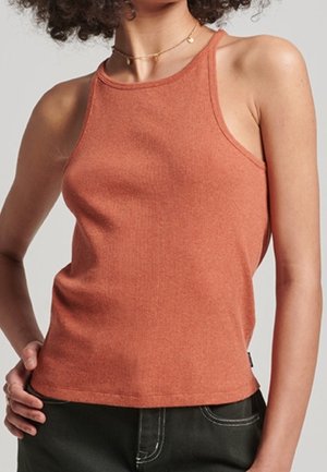 Mujer con una camiseta sin mangas de color óxido combinada con pantalones oscuros, de pie con una mano en la cadera frente a un fondo liso.