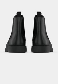 Pár černých kožených kotníkových bot zobrazených ze zadní strany na jednoduchém bílém pozadí, s poutky a silnými gumovými podrážkami.