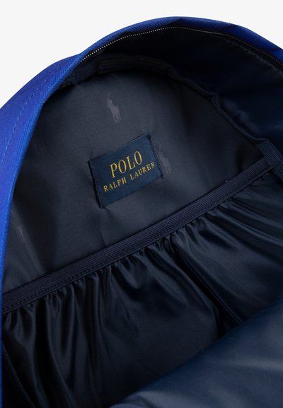 Interior de una bolsa de tela azul que presenta un forro azul marino, una etiqueta de POLO RALPH LAUREN y bolsillos elásticos a lo largo de los bordes.
