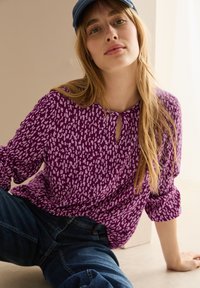 Top viola a maniche lunghe con scollatura a chiave, caratterizzato da un motivo texturizzato in viola più chiaro. Abbinato a jeans in denim scuri.