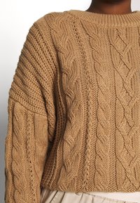 Cable-knit sweater in tan, featuring ribbed details, a rounded neckline, and chunky texture. Relaxed fit with wide sleeves.