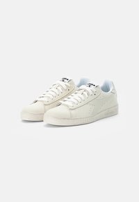 Baskets en cuir blanc avec des perforations, lacets plats et semelle en caoutchouc texturée. Présentent un logo sur la languette et des détails de couture subtils.