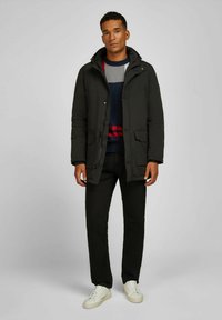 Jonge man in een zwarte gewatteerde winterjas, met een gebreid patroon op zijn trui, zwarte broek en witte sneakers, staande voor een effen lichtgrijze achtergrond.